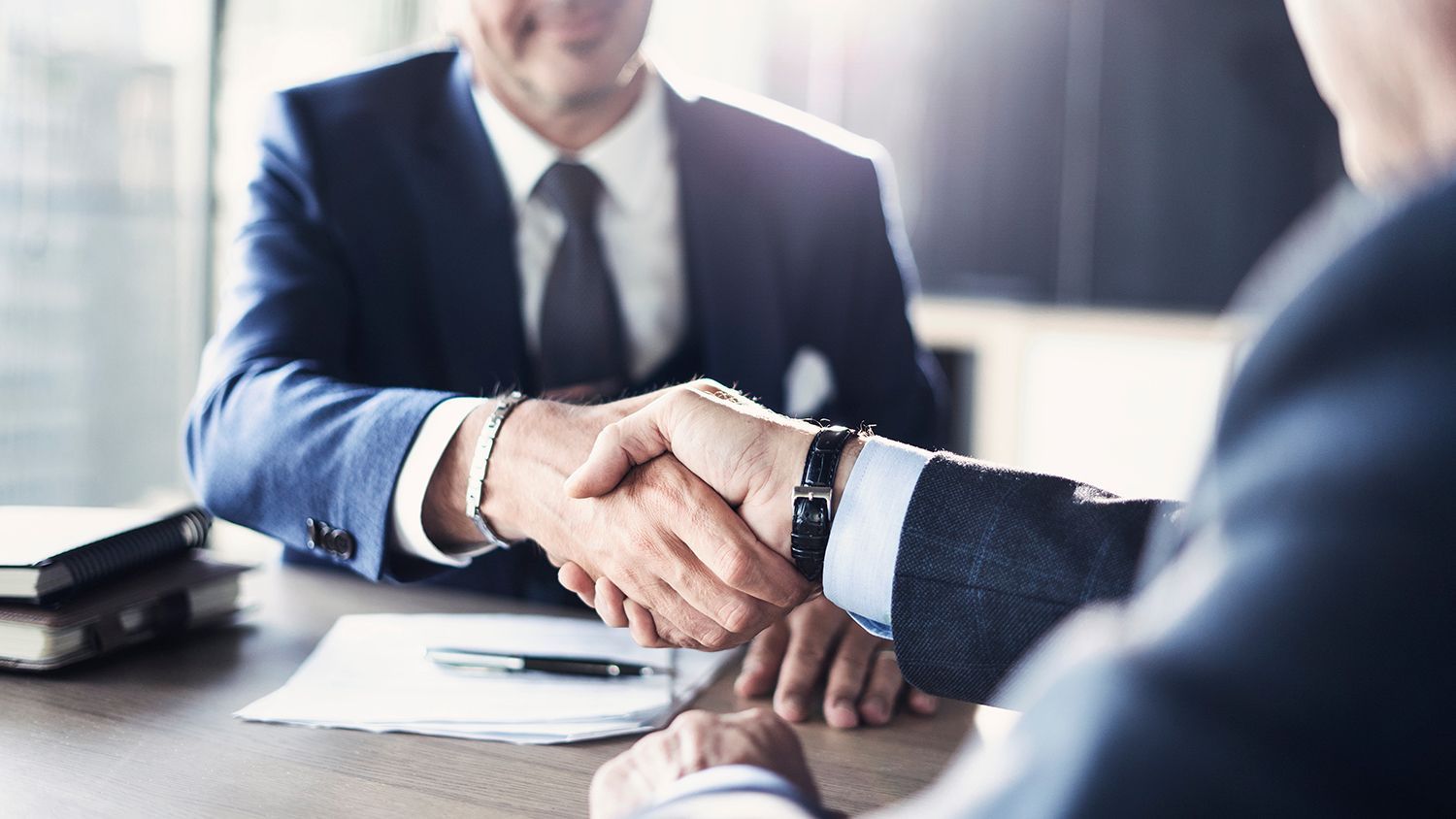 Two businesspeople shake hands across a desk in a bright office Two businesspeople shake hands across a desk in a bright office