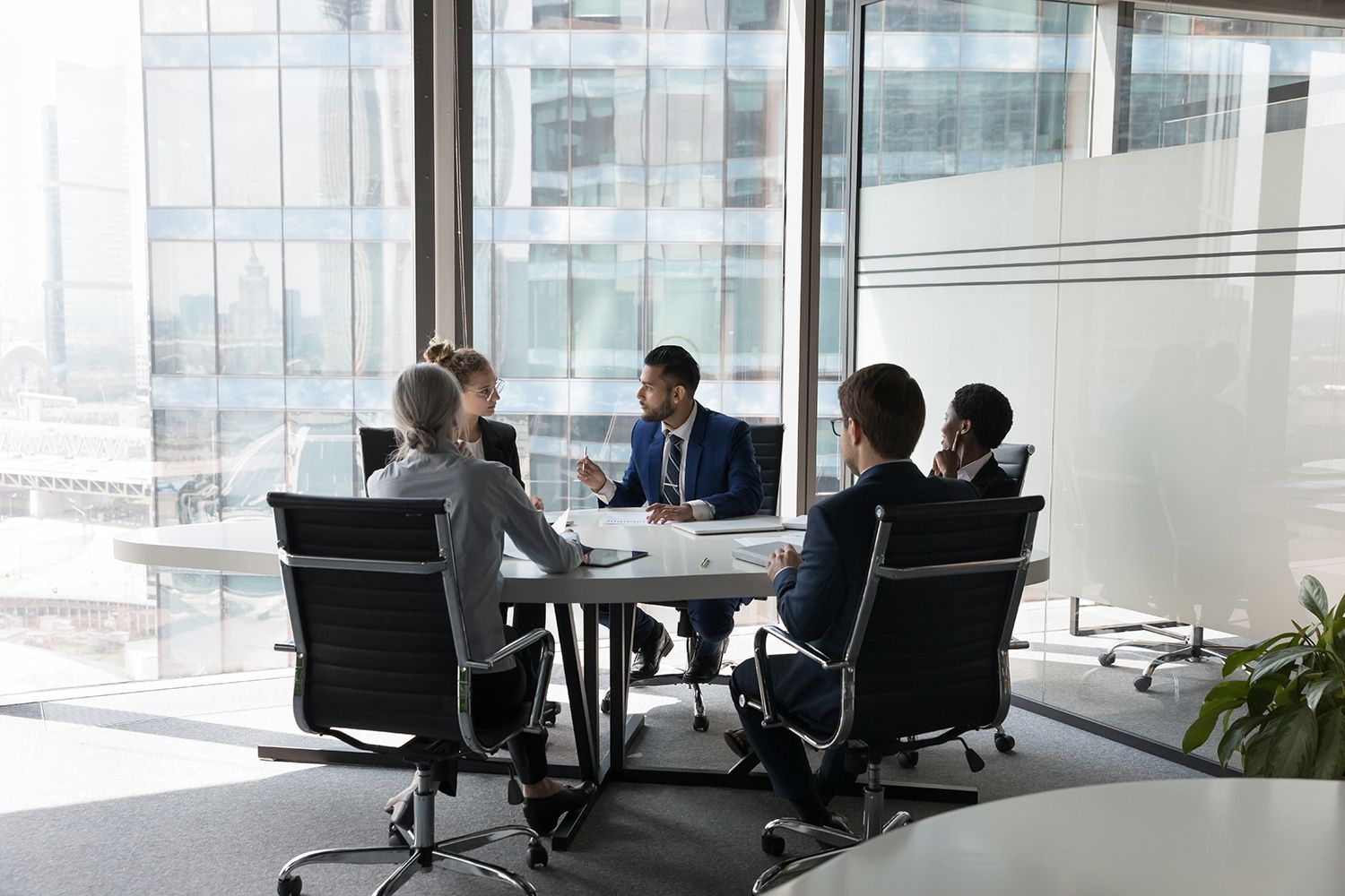 Business meeting in a bright glass-walled conference room, with four people seated around a round table. Business meeting in a bright glass-walled conference room, with four people seated around a round table.