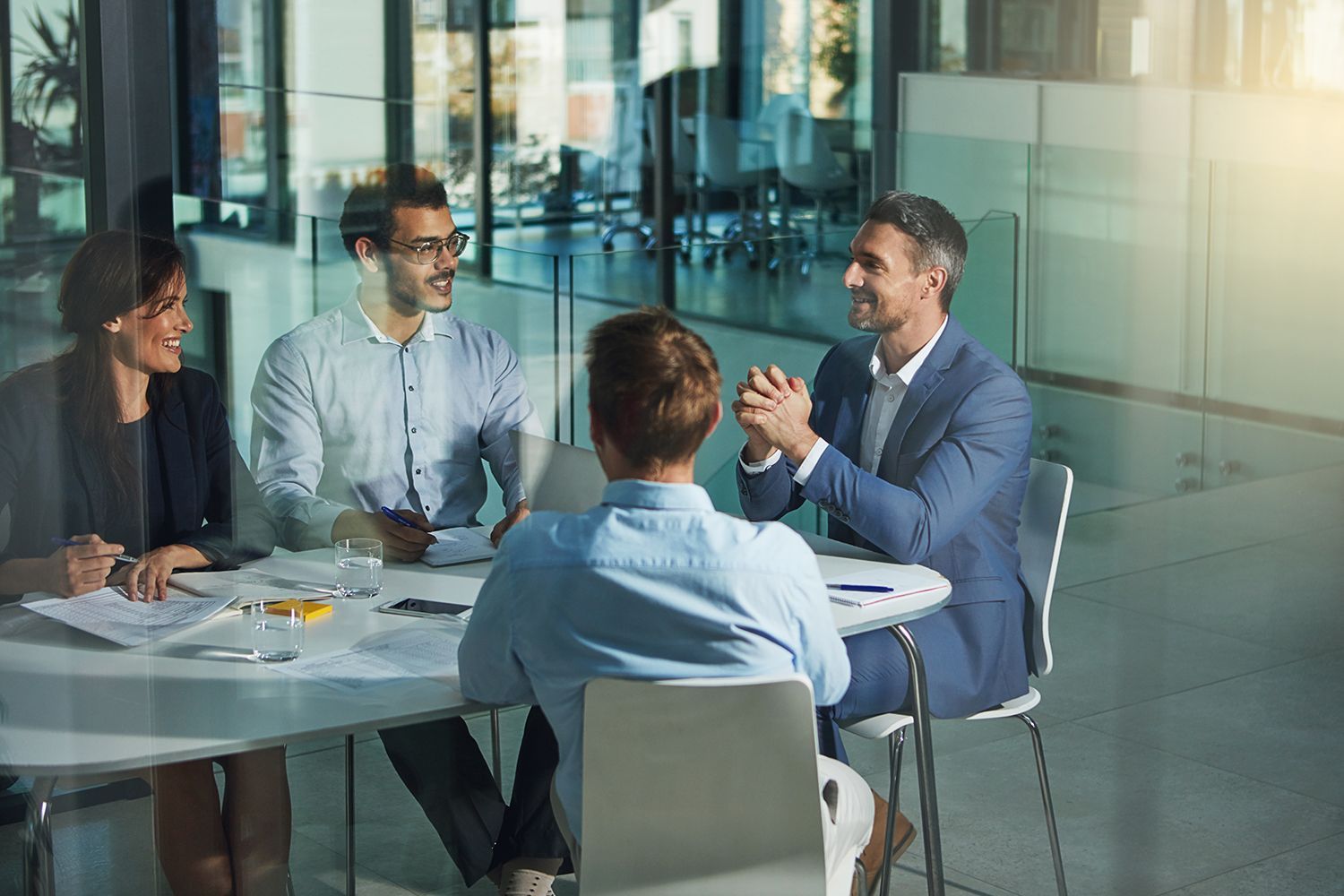 Four coworkers in a glass conference room discussing around a table Four coworkers in a glass conference room discussing around a table