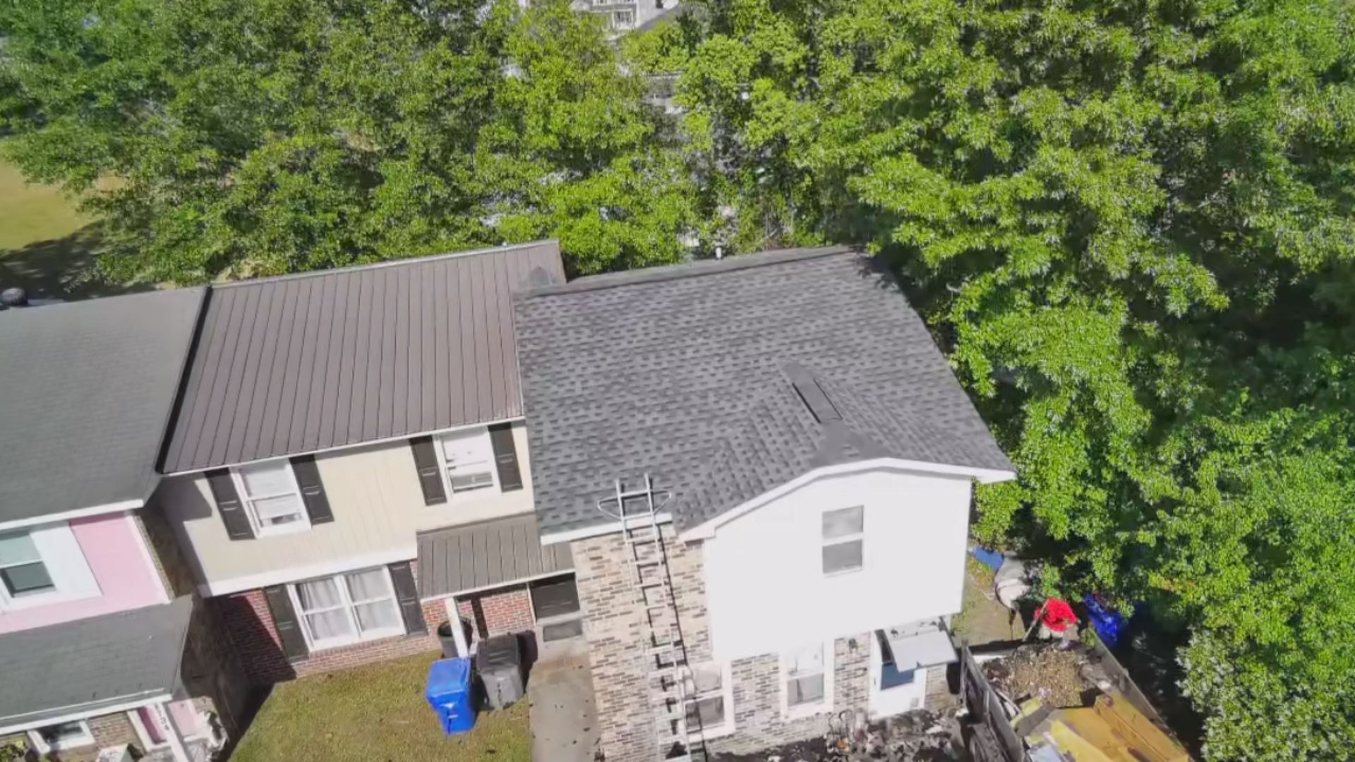 Aerial view of two attached houses with gray roofs and trees around them, with a small deck and yard below.