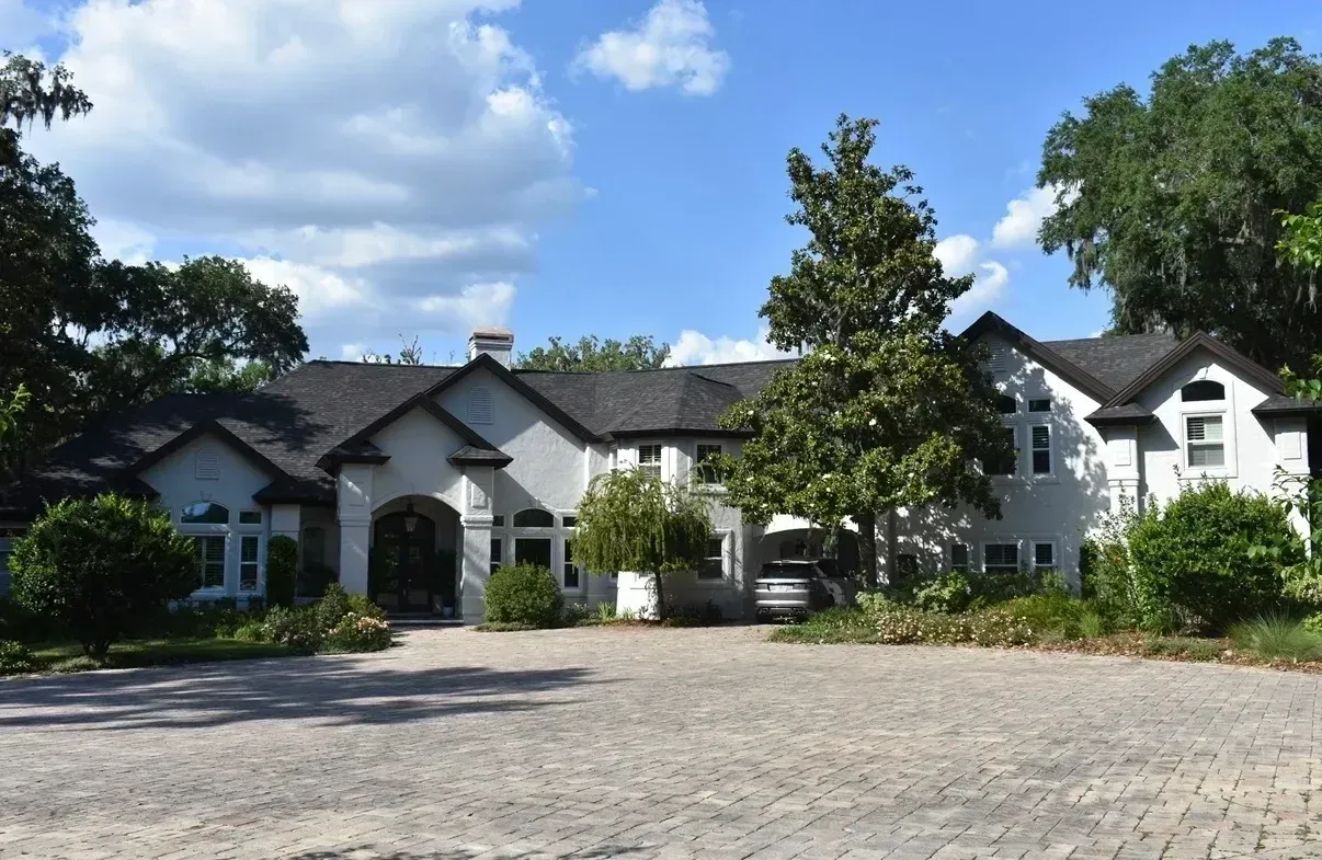 White house with dark roof and trees under a blue sky, viewed from a cobblestone driveway