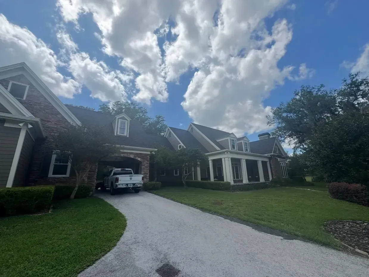 Large brick house with a gravel driveway, white carport, and green lawn under a cloudy sky
