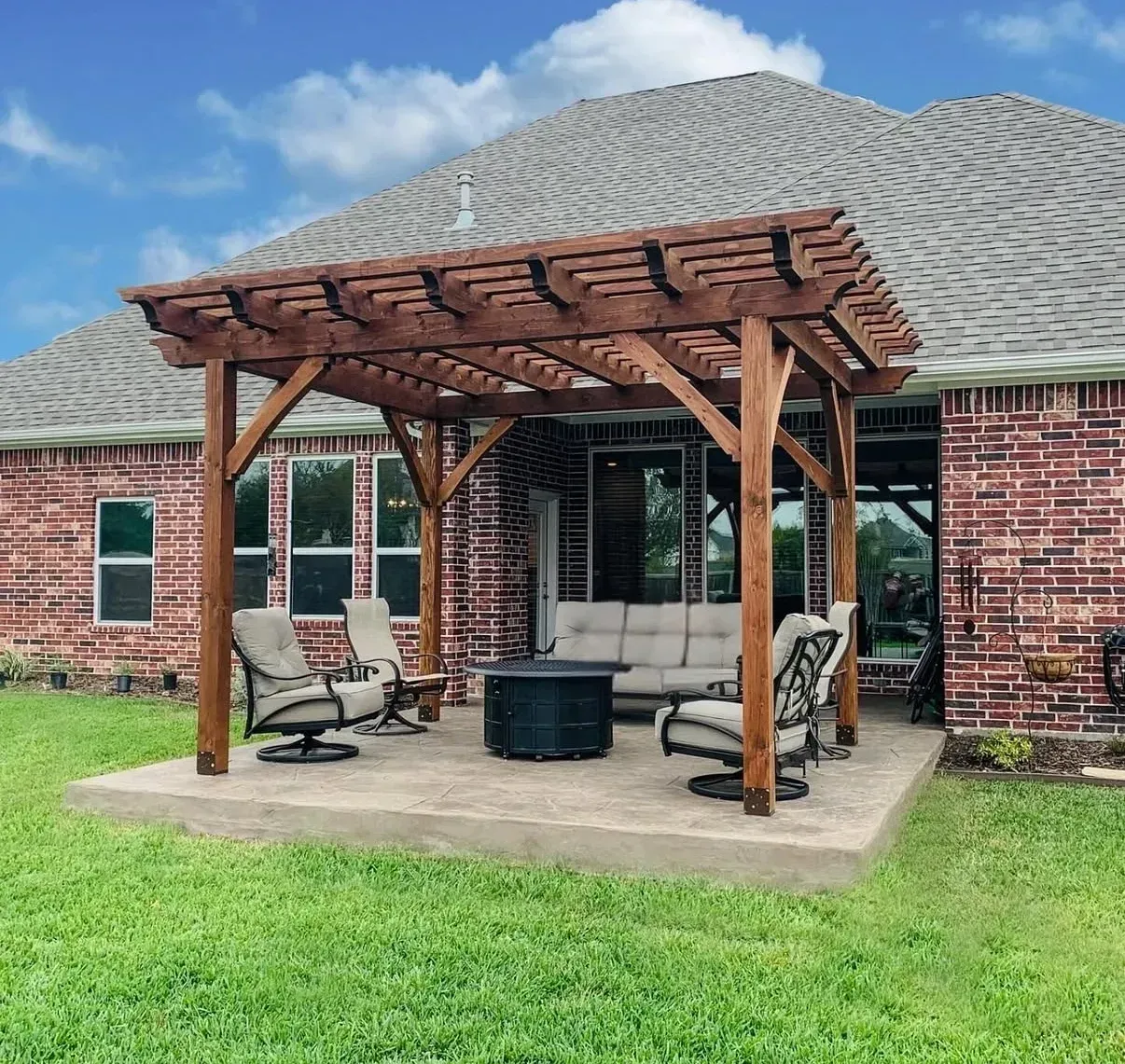 Backyard patio with wooden pergola, seating, and fire pit beside a brick house.