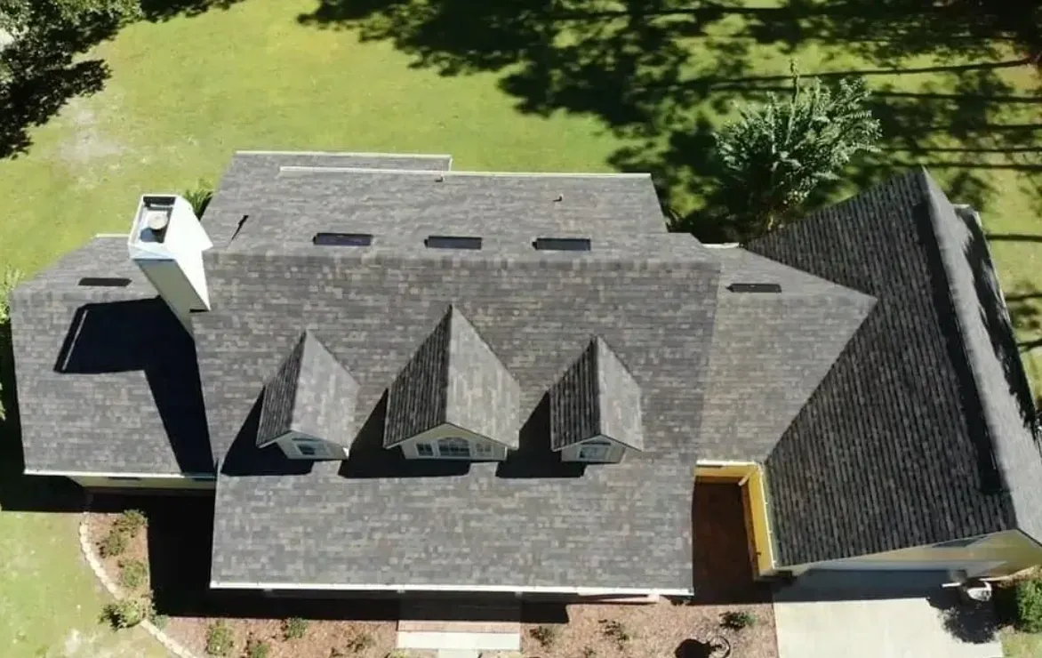 Aerial view of a gray-shingled house roof with dormers, chimney, and surrounding green lawn.