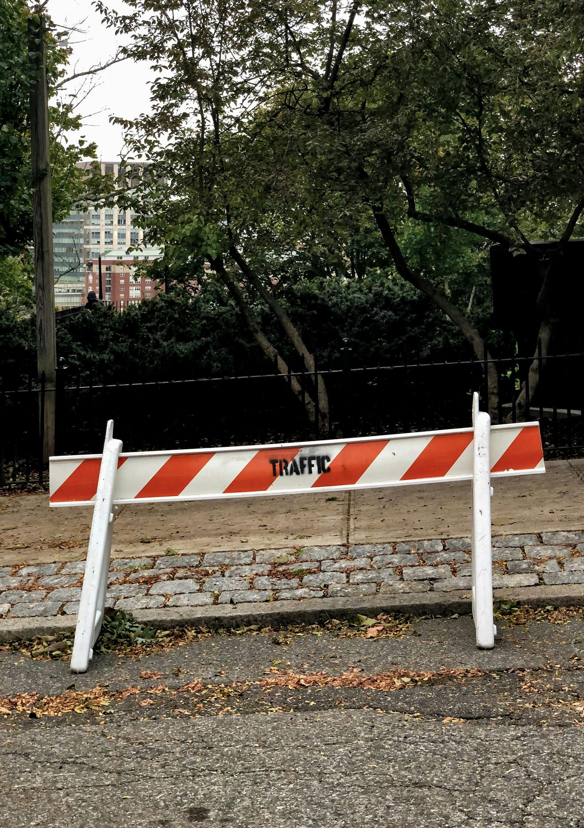 White and orange striped traffic barricade on a city street with foliage in the background. 