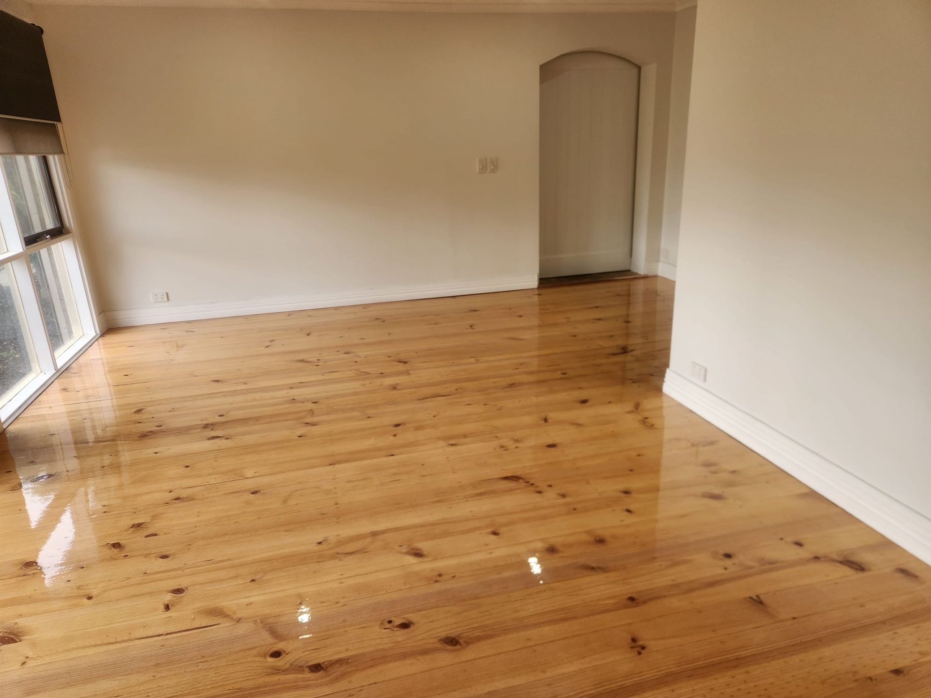 Empty room with polished wooden floor, white walls, window blinds, and an arched doorway.
