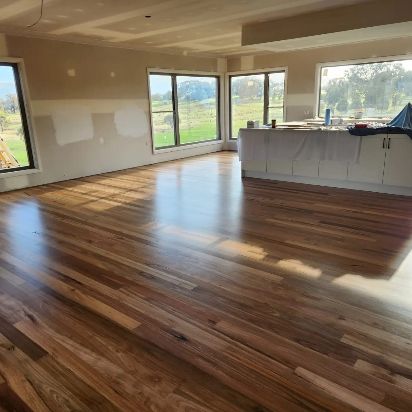 Bright empty room with polished wood floors, large windows, and a kitchen island overlooking green fields