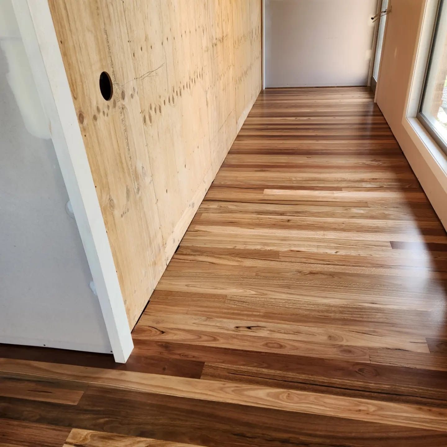 Narrow hallway with striped wood flooring, pale walls, and a window on the right.