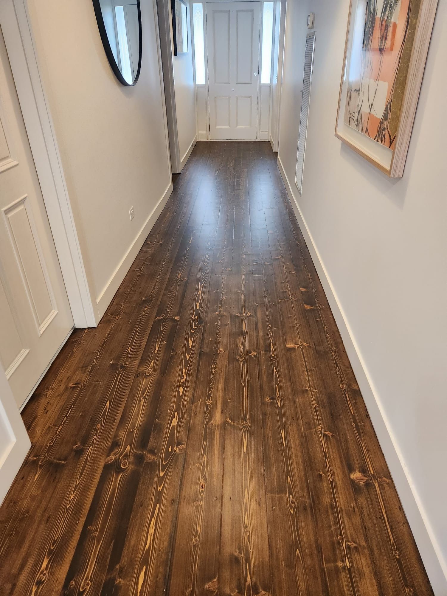 Narrow hallway with dark wood floors, white walls, and framed artwork leading to a white front door