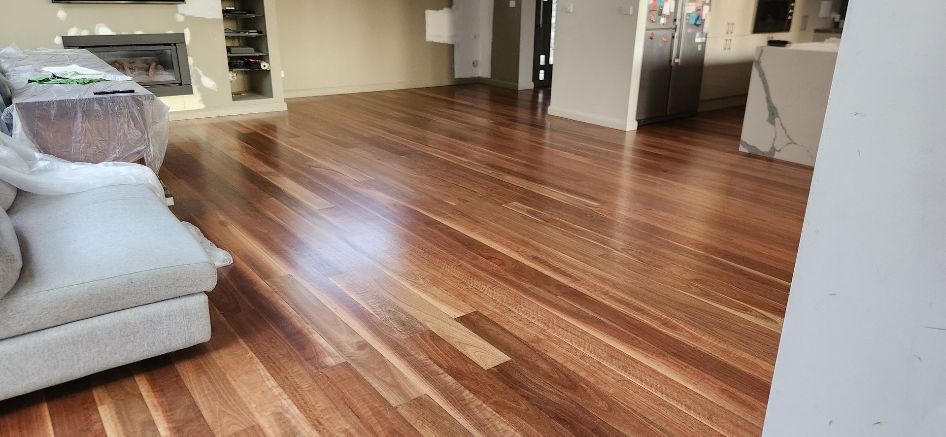 Empty room with polished hardwood floors, a light gray sofa, and kitchen area in the background