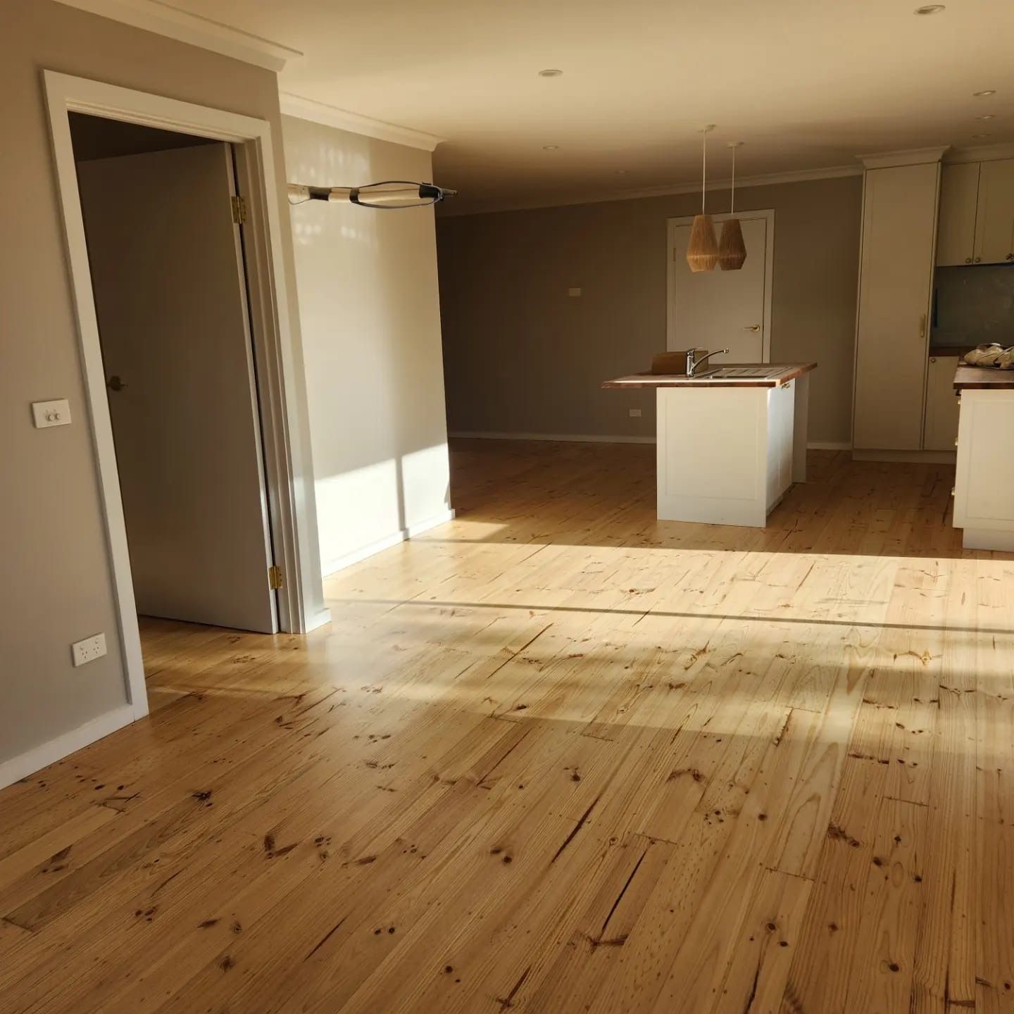Empty sunlit living room with light wood floor and open kitchen area