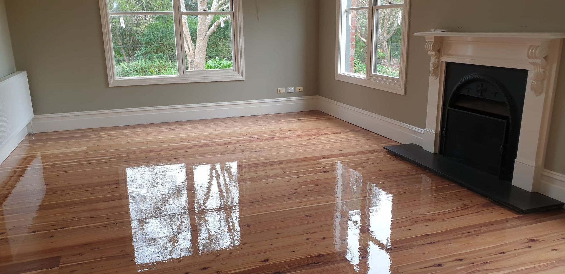 Empty room with glossy hardwood floors, beige walls, large windows, and a black fireplace.