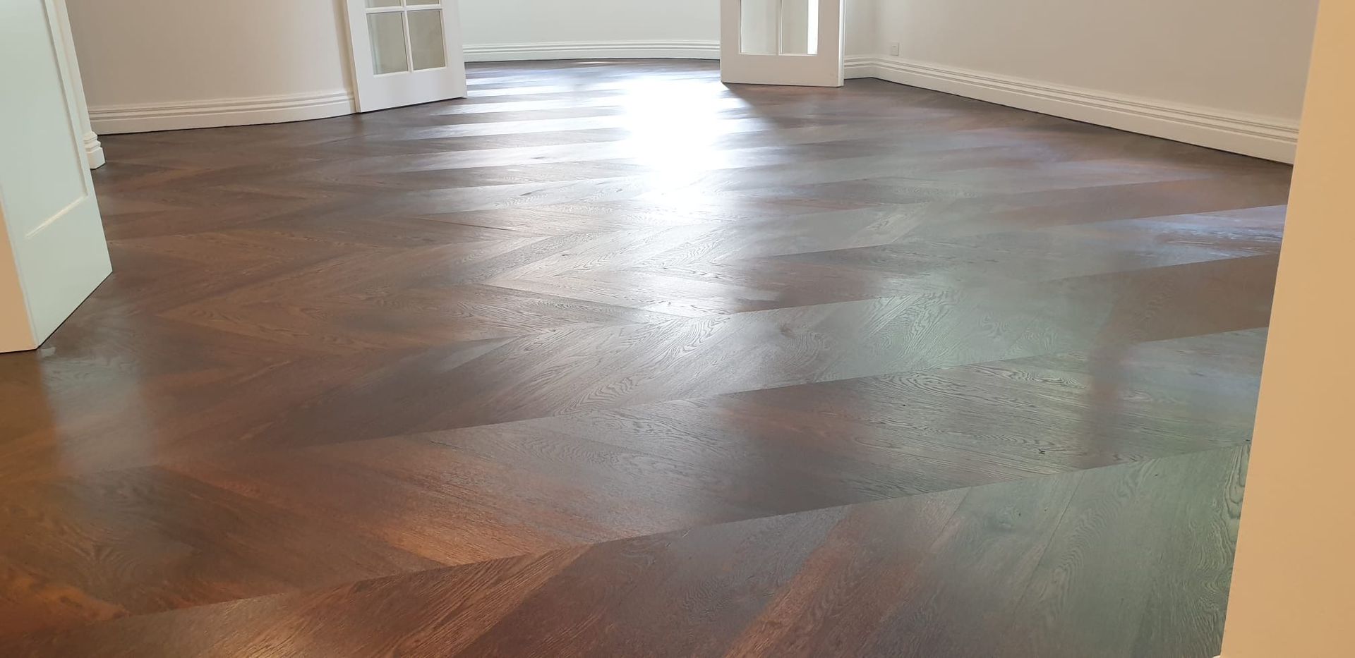 Empty room with dark herringbone wood floor and white walls, lit by natural light.