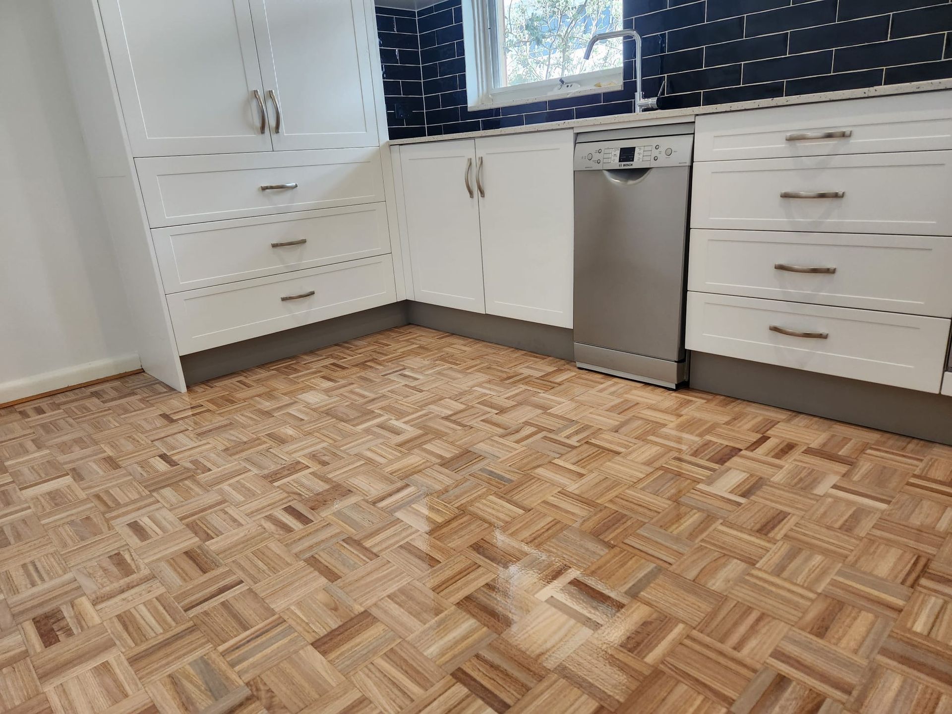 Empty kitchen with white cabinets, stainless dishwasher, and herringbone wood floor.