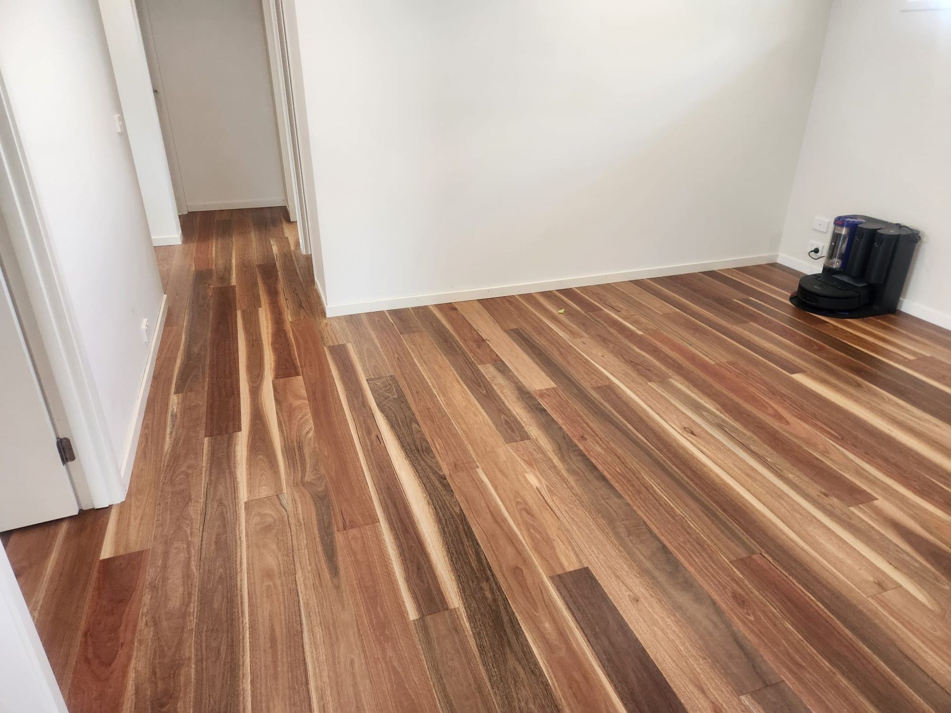 Empty room with striped wood flooring, white walls, and a black appliance in the corner