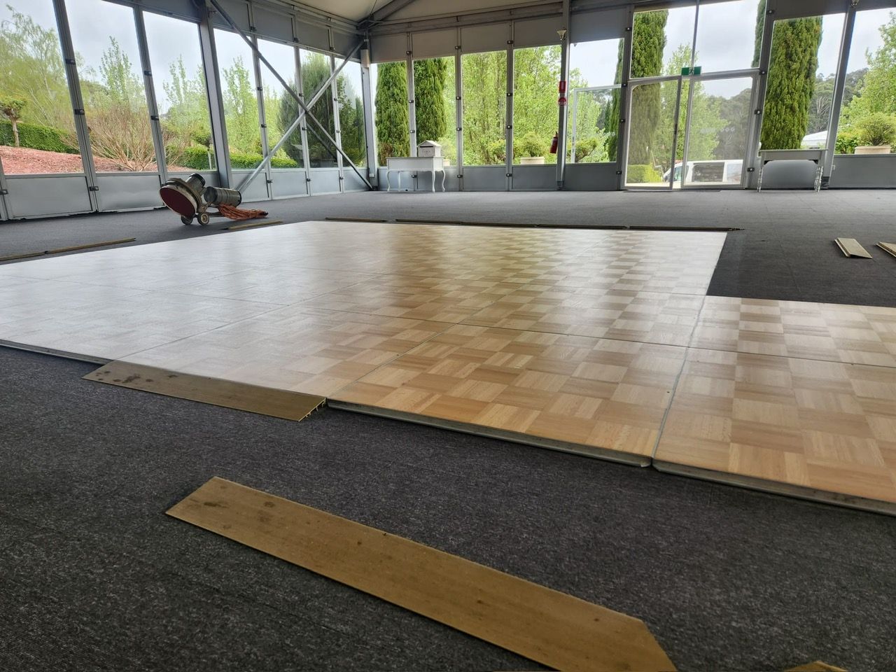 Indoor hall with a polished wooden dance floor, gray carpet, and large windows overlooking greenery.