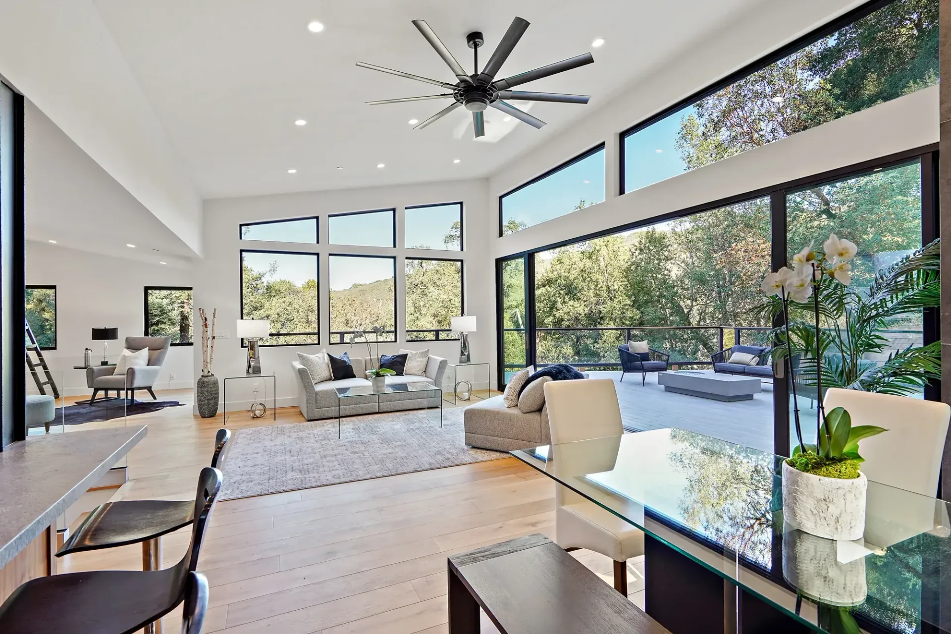 Modern living room with large windows overlooking a deck and trees. Bright, airy space with neutral tones.