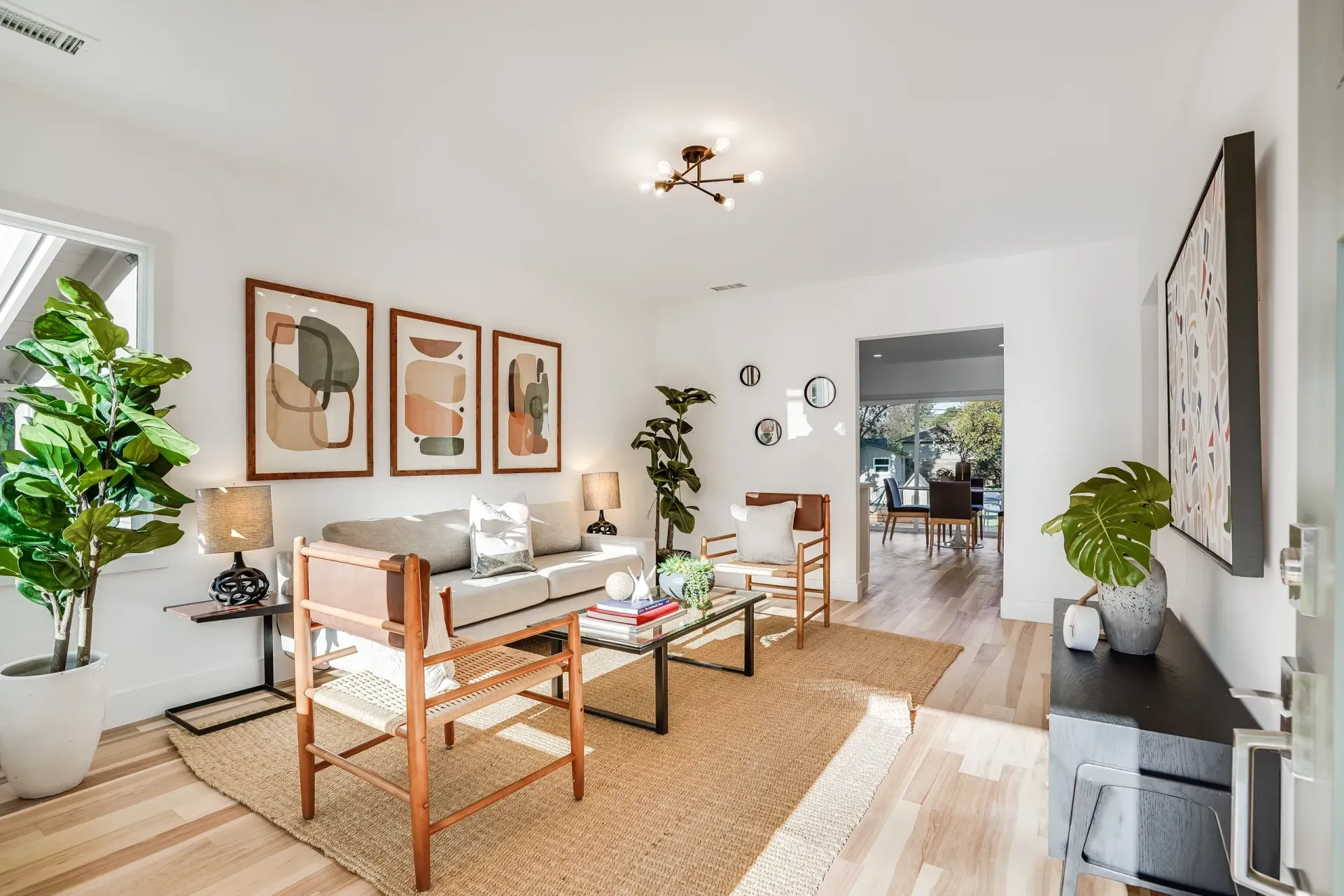 Cozy living room with neutral tones, three framed prints, and a jute rug. Features a sofa, chairs, and potted plants.