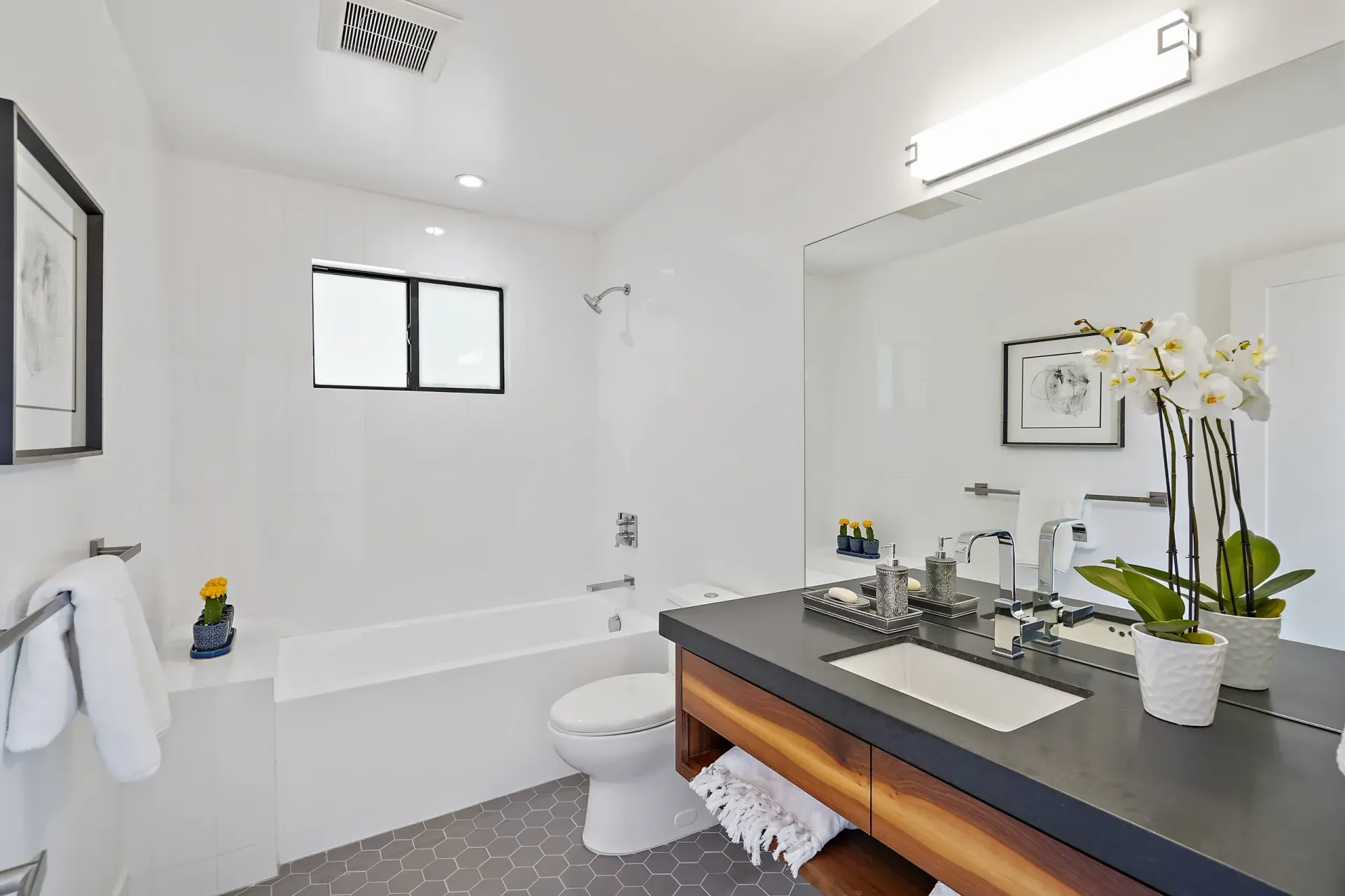 Modern white bathroom with a tub, toilet, large mirror, and orchids on the countertop.
