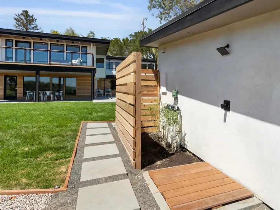Outdoor shower with wooden screen and walkway beside a modern house.