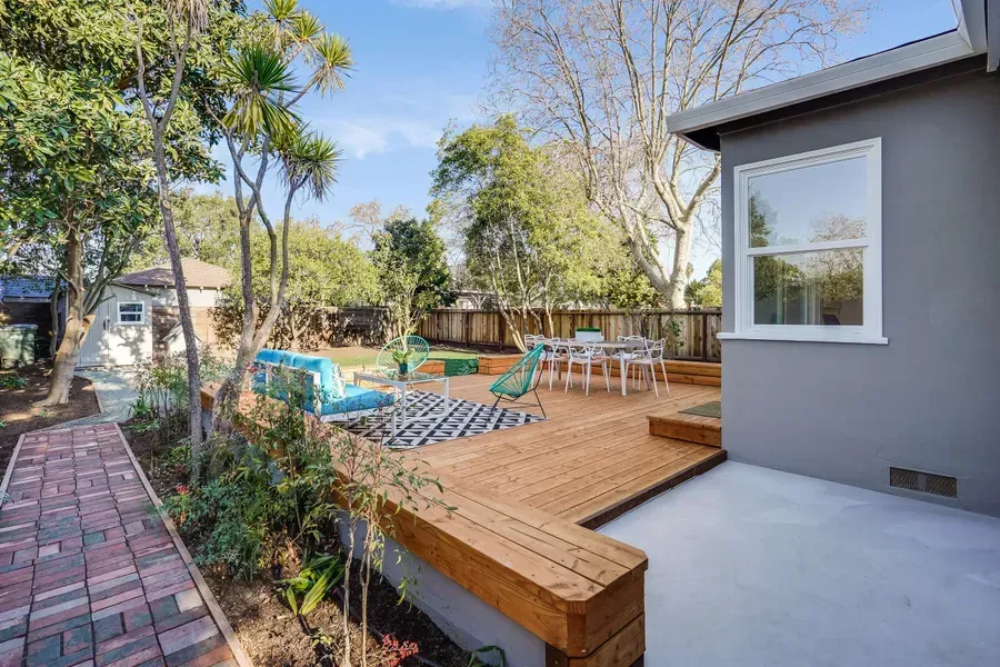 Wooden deck with outdoor furniture, and brick pathway. Lush backyard with trees, blue skies.