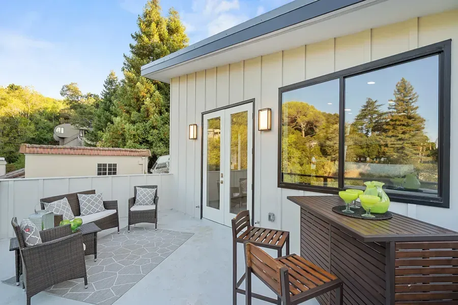 Outdoor deck with seating, bar, and door to a home, overlooking trees.