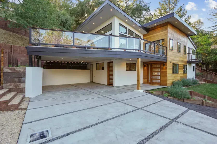 Modern two-story house with a flat roof, wood siding, and glass balcony overlooking a concrete driveway.