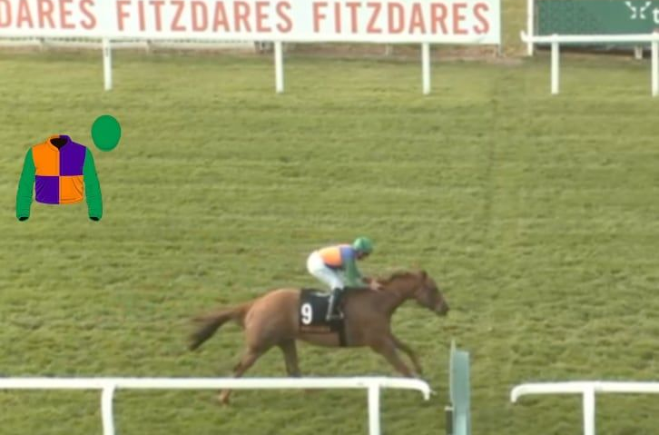 Horses racing on a green track, jockey in blue leading, others close behind, overcast day, near white fence.