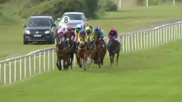 Horses racing on a green track, jockey colors include yellow, pink, and blue. A white fence and car are in the background.