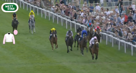 Jockeys race horses down a grassy track with spectators behind the rail.
