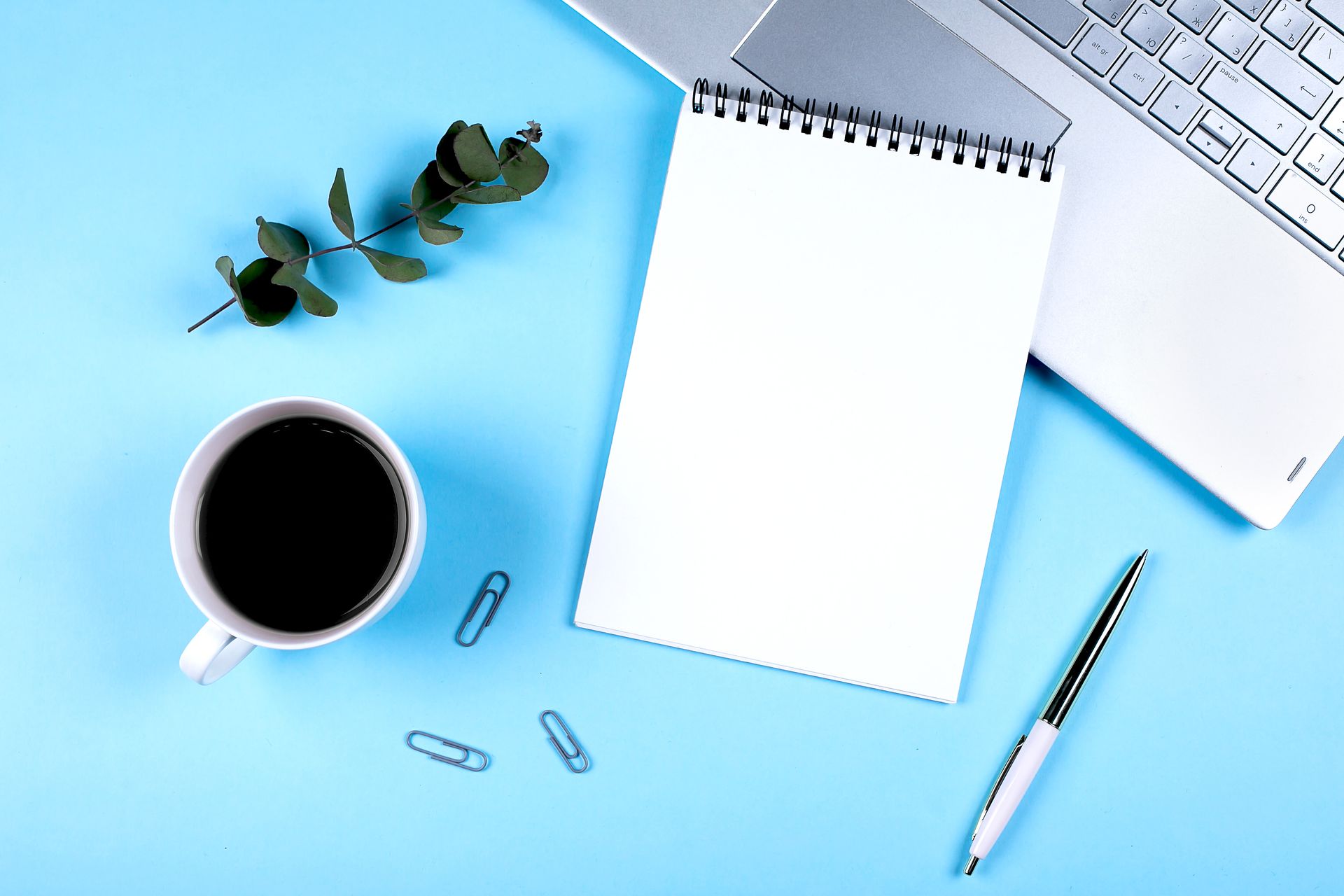 A cup of coffee , a notebook , a pen , and a laptop on a blue table.