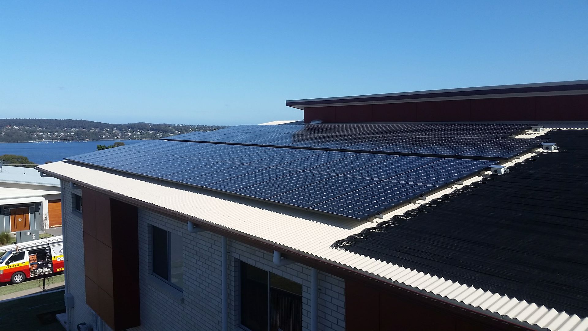 Rooftop covered with solar panels beside a waterfront, with blue sky and distant buildings— Solar Services in Erina, NSW