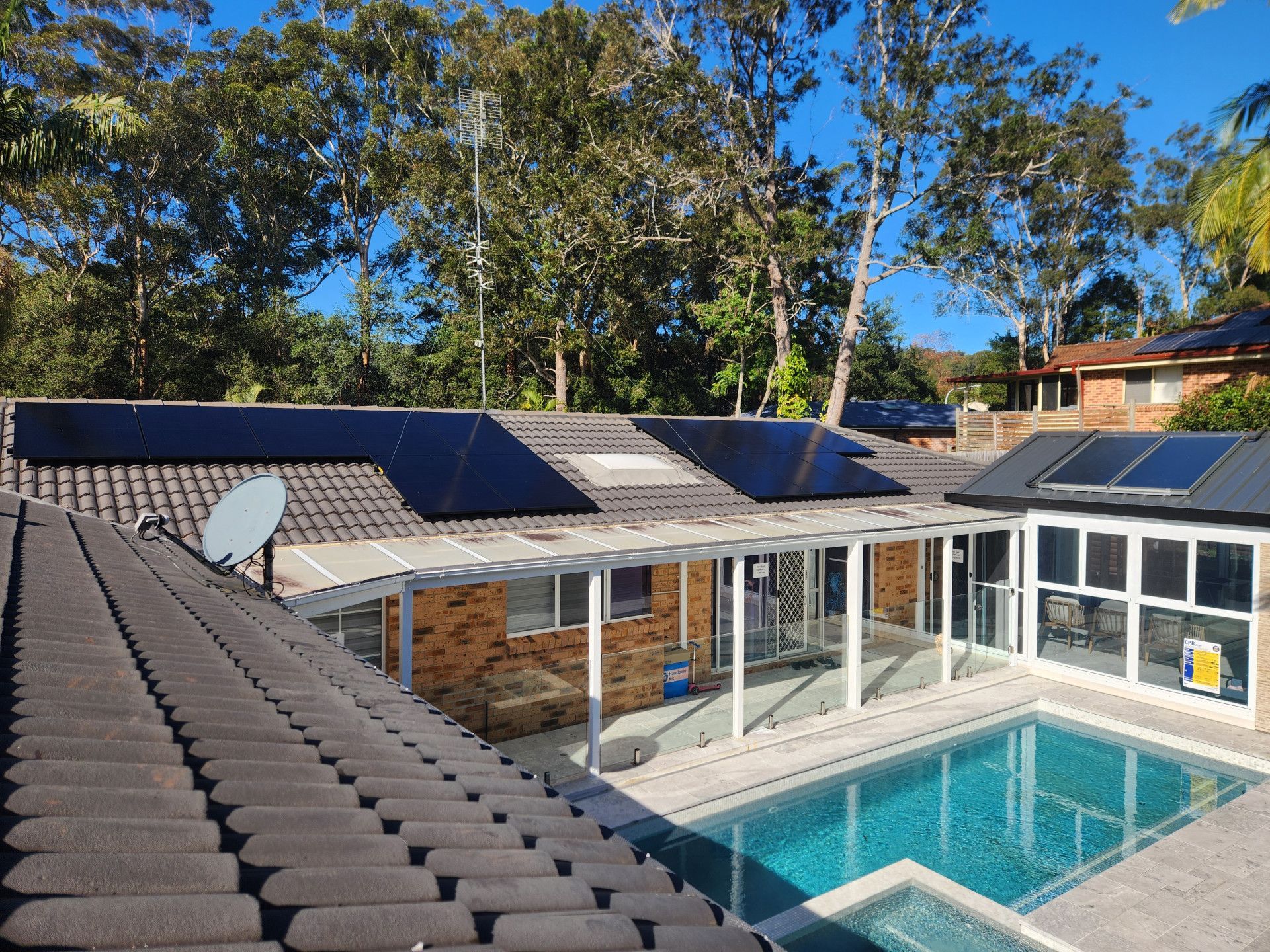 Rooftop solar panels beside a turquoise swimming pool and glass-enclosed patio in a sunny yard — Solar Services in Erina, NSW