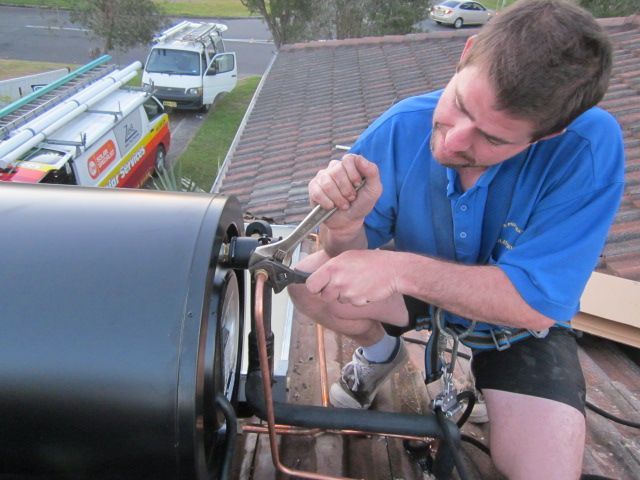 Man on a roof using a wrench on a black water heater. He is wearing blue shirt, shorts, and safety harness  — Solar Services in Erina, NSW