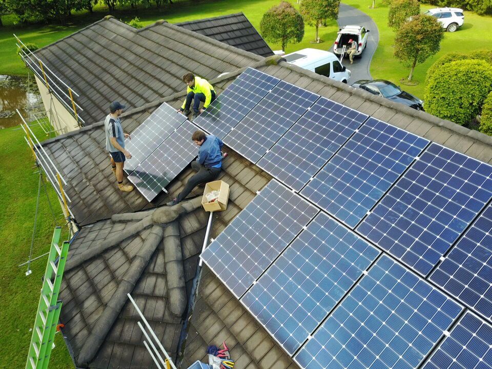 Workers installing solar panels on a house roof with black shingles and green lawn nearby — Solar Services in Erina, NSW