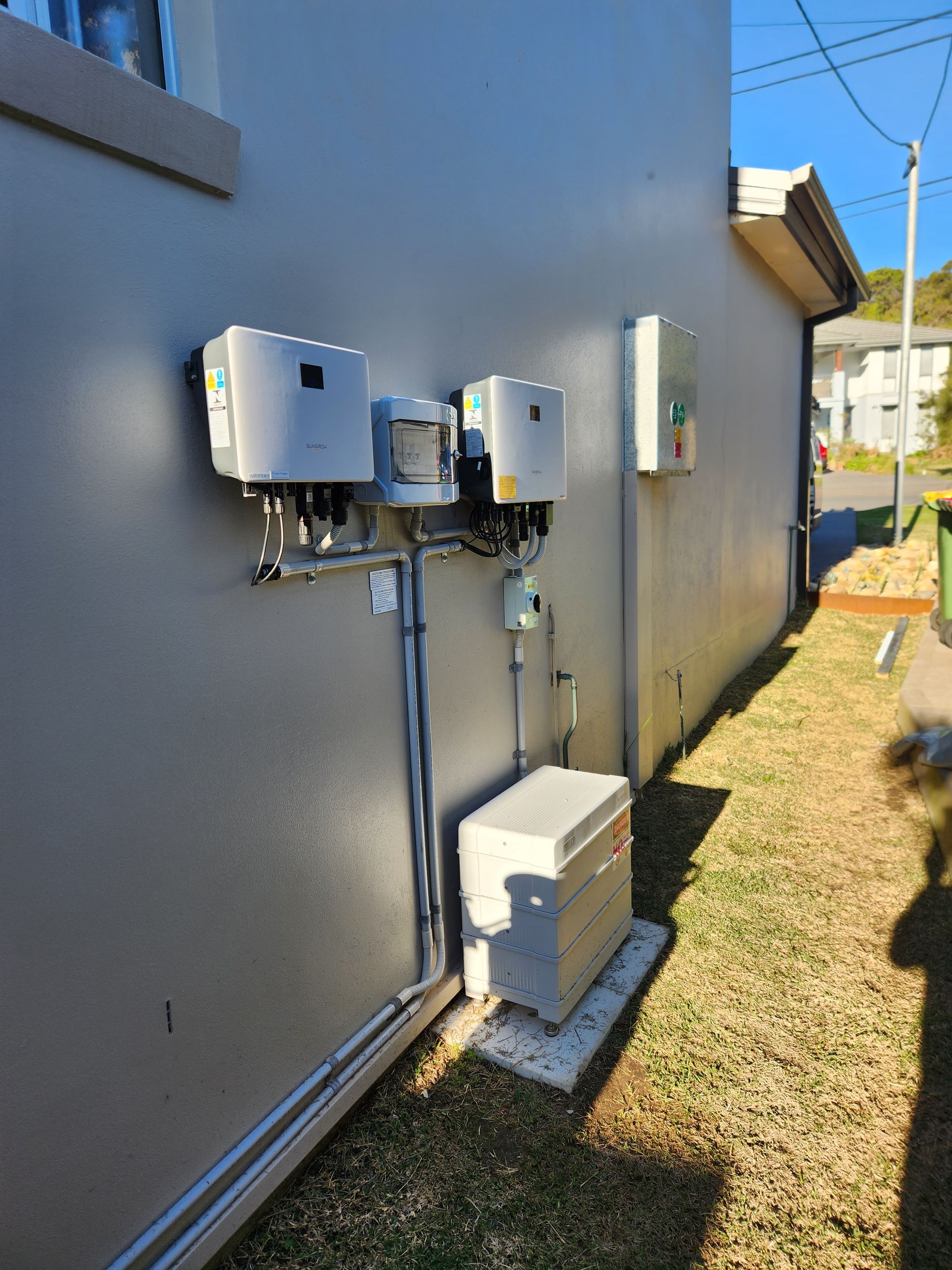 Outdoor Solar Battery inverters mounted on a house wall beside a grassy yard — Solar Services in Erina, NSW