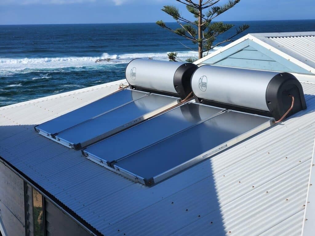 Rooftop solar water heater with blue collectors on a white house beside the ocean — Solar Services in Erina, NSW