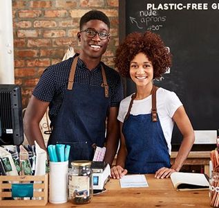 Two smiling café workers in blue aprons behind a counter with reusable cups and a chalkboard sign