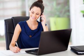 Woman on the phone, smiling at a laptop while writing notes at a desk