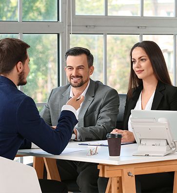 Three people in a bright office meeting around a table with a laptop and coffee cup.