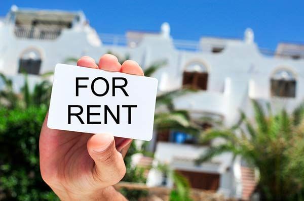 Hand holding “FOR RENT” sign in front of a white house with palm trees