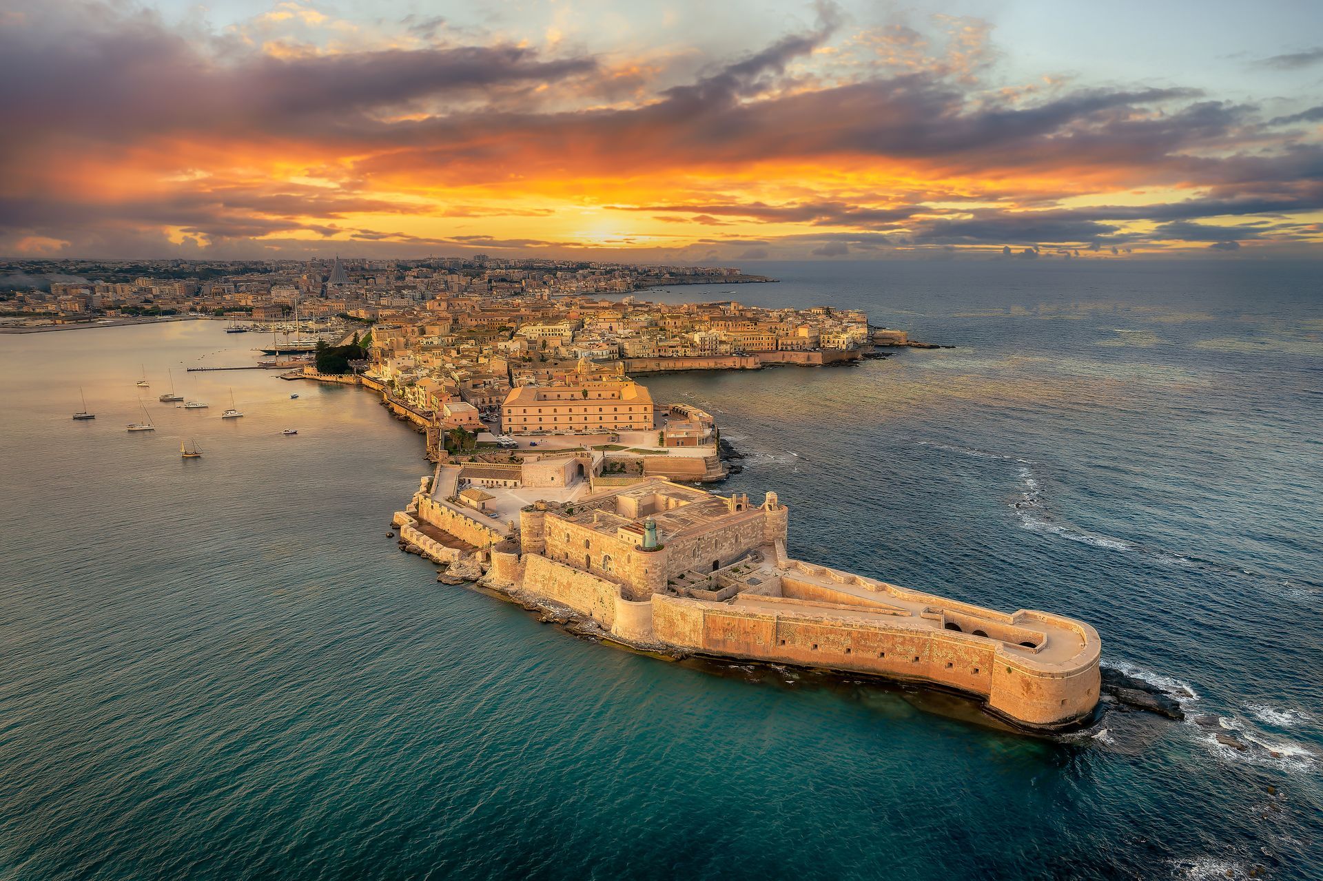 Vista aerea di una fortezza costiera nel mare al tramonto, con sullo sfondo una città e un cielo arancione.