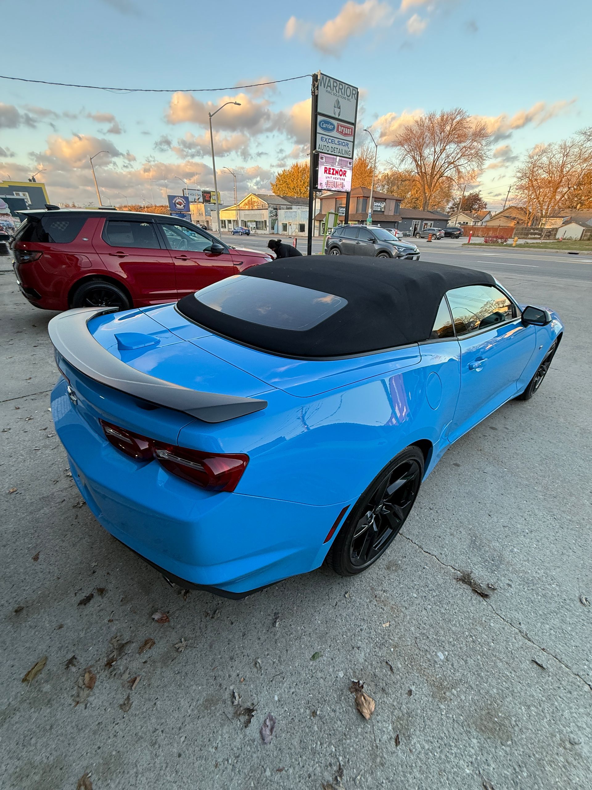 Blue convertible sports car parked in a lot at sunset, with a red SUV nearby.
