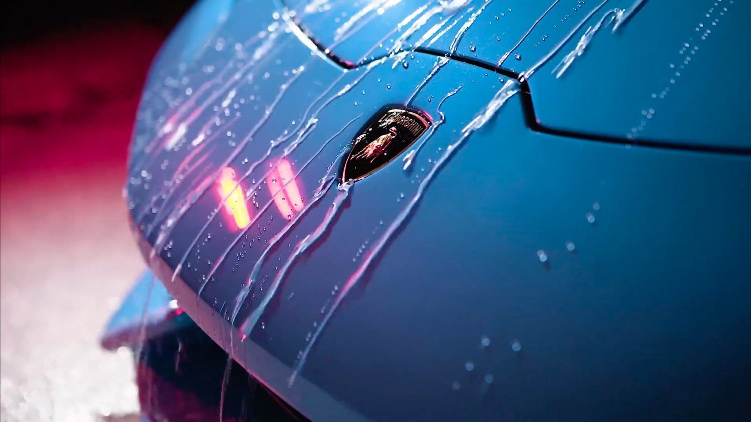 Close-up of a blue car’s front end with water droplets and pink reflections in the wet paint