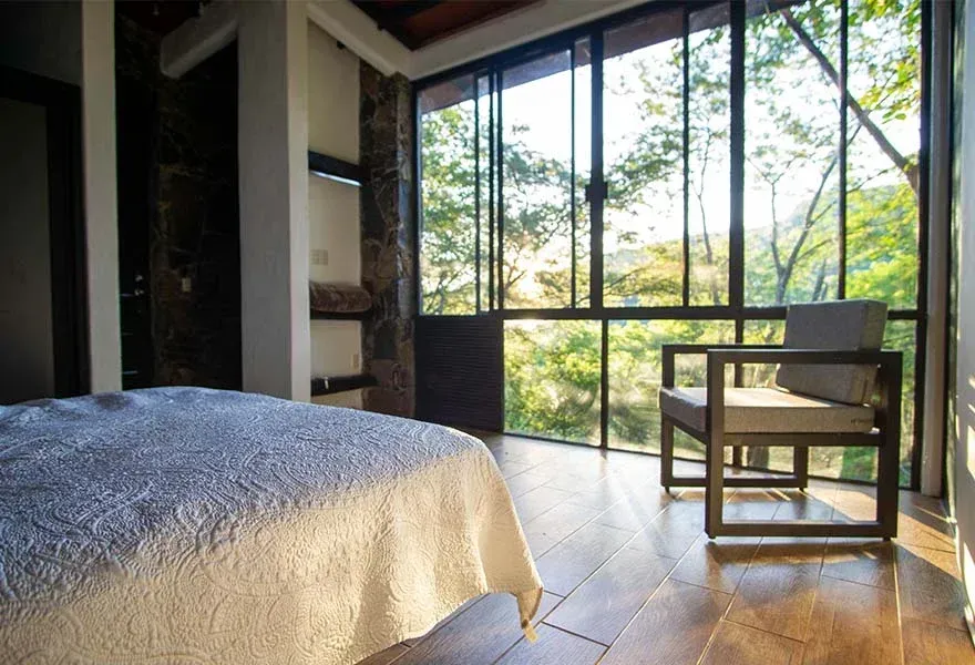 Habitación luminosa con cama, silla de madera y grandes ventanales con vistas a la vegetación.
