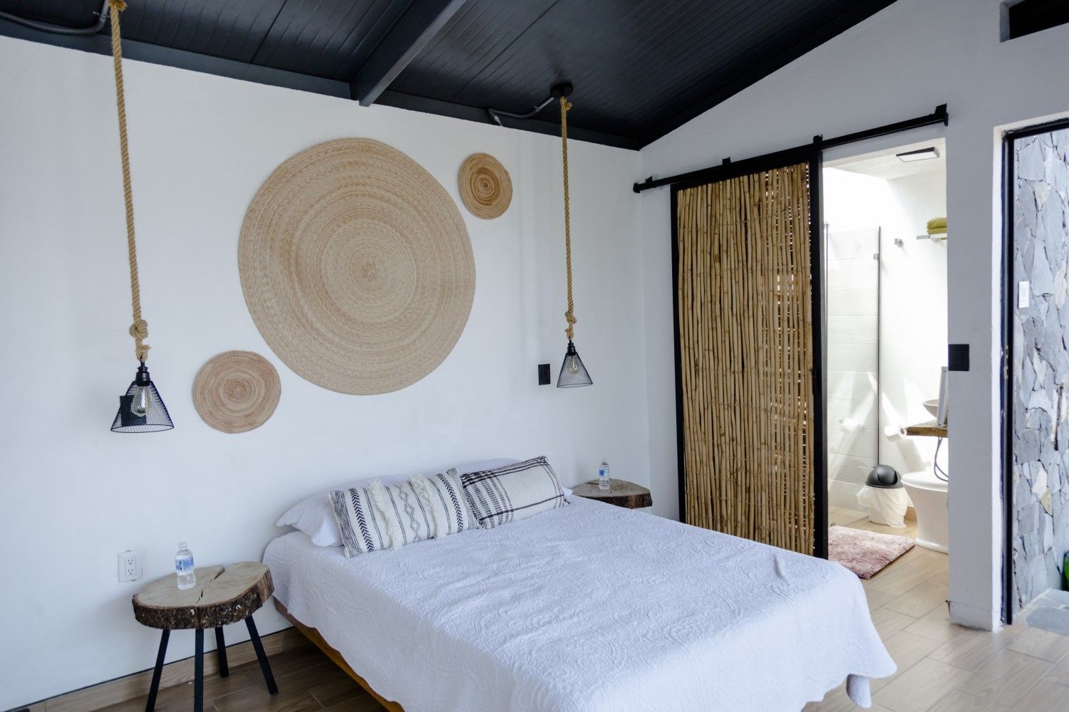 Dormitorio luminoso y minimalista con cama blanca, decoración de pared tejida, lámparas colgantes y puerta abierta.