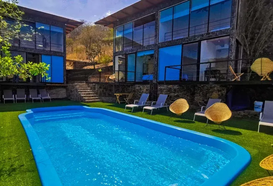 Patio de una casa moderna con una piscina de color azul brillante, tumbonas y grandes ventanales.
