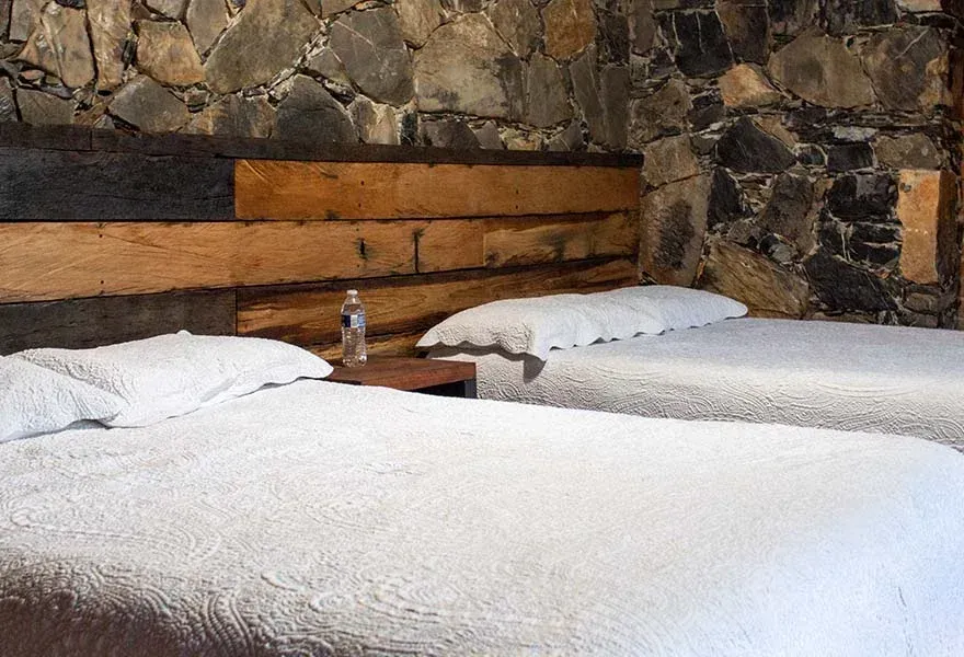 Dos camas blancas con cabecero de madera contra una pared de piedra en una habitación rústica.