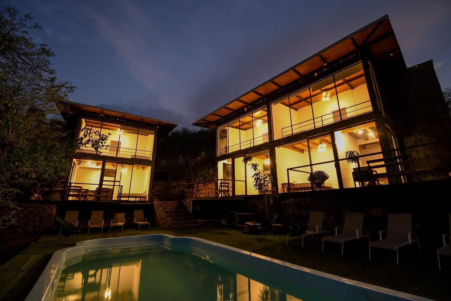 Villa moderna de cristal que brilla por la noche junto a una piscina iluminada.