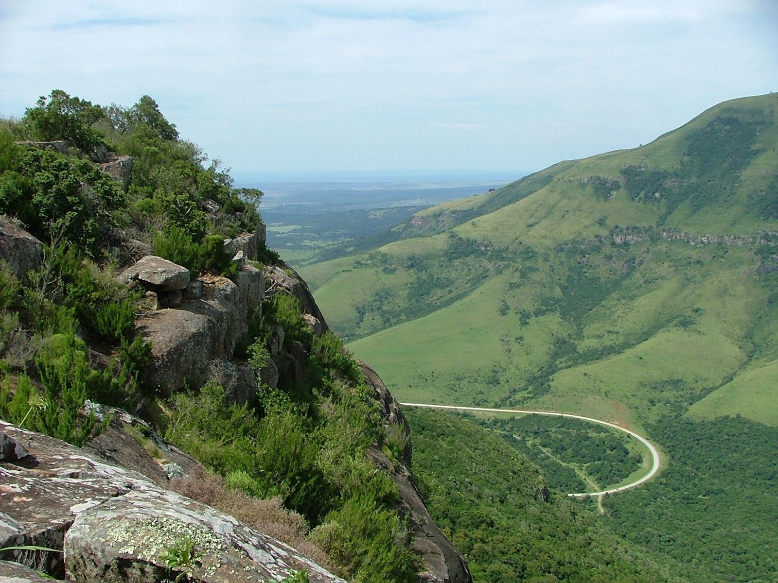 Green mountains and valley with a winding road; rocky cliff in foreground.